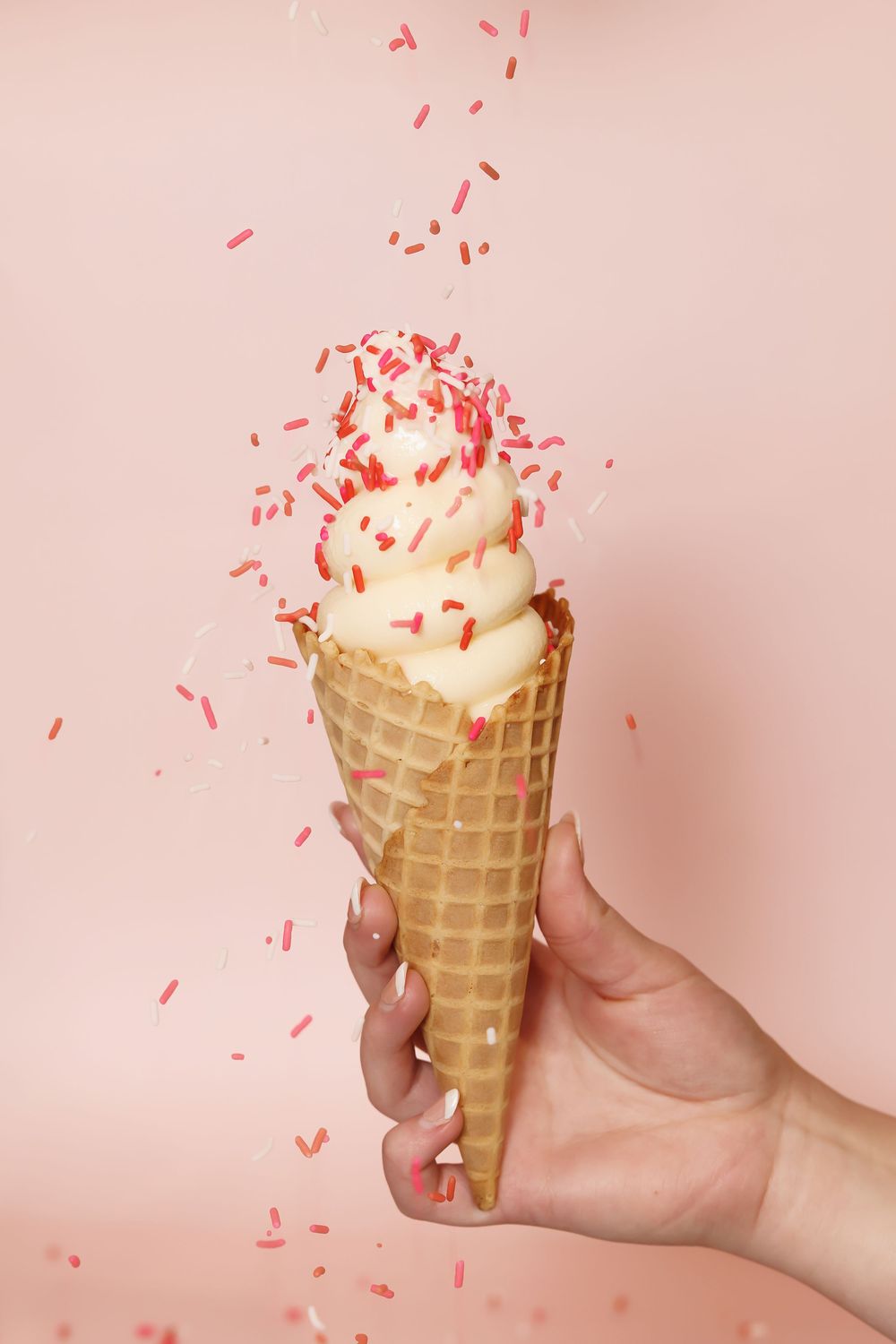 Ice cream cone topped with swirled vanilla soft serve and red sprinkles being held against a pink background.
