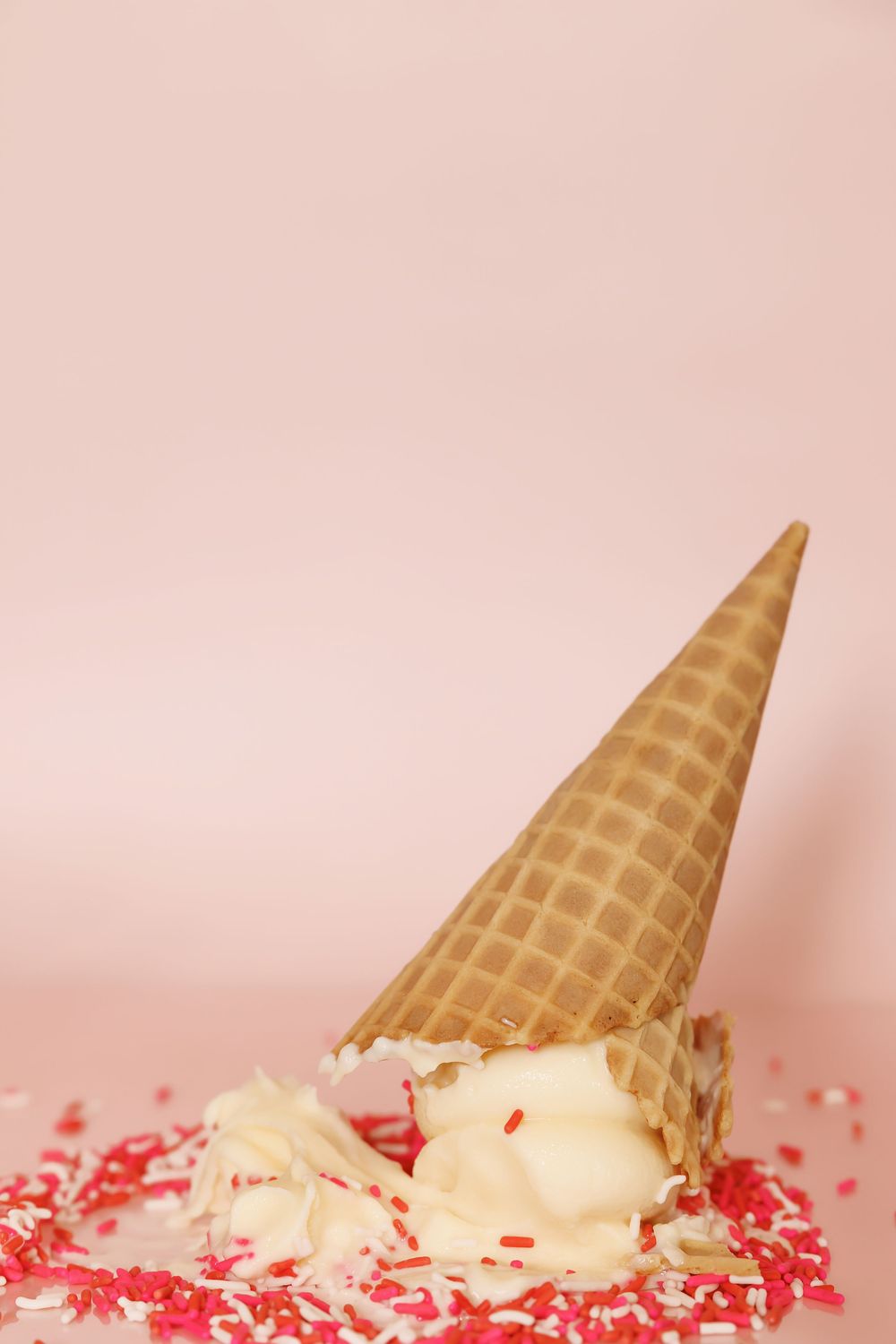 Fallen waffle cone with vanilla ice cream and pink sprinkles against a pale pink background.