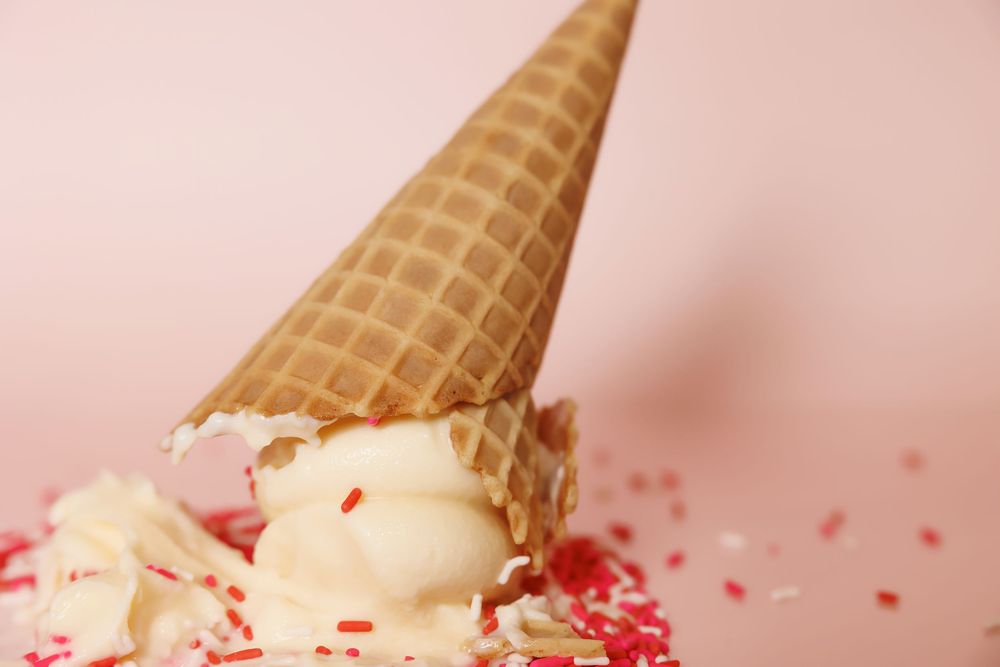 A tipped over waffle cone with vanilla ice cream and red candy sprinkles scattered against a pink background.