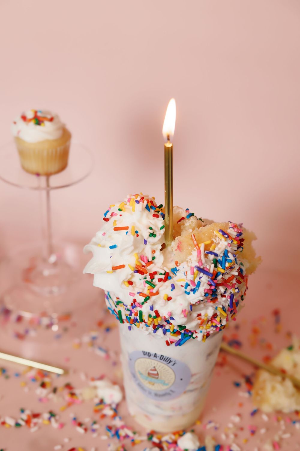 Birthday milkshake decorated with sprinkles and lit candle on pink background with cupcake in soft focus.