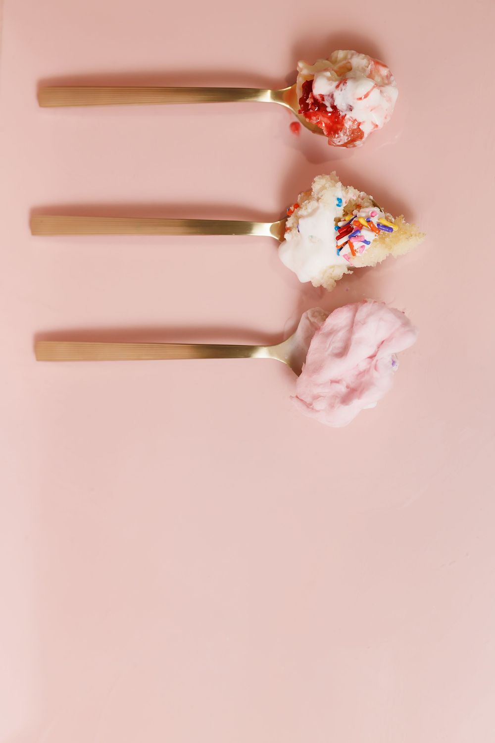 White plastic spoons topped with pink ice cream and colorful sprinkles on a pink surface.