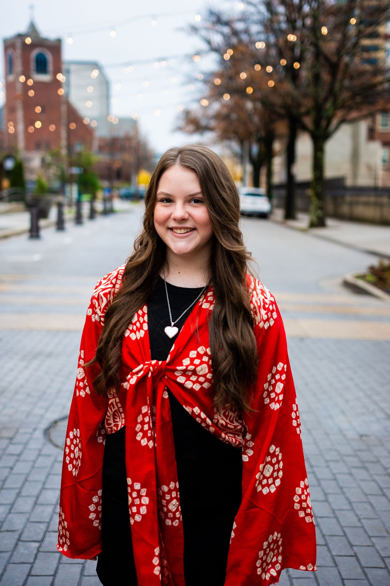 Person standing on urban sidewalk wearing bright red floral kimono wrap over black dress in downtown setting with twinkle lights.