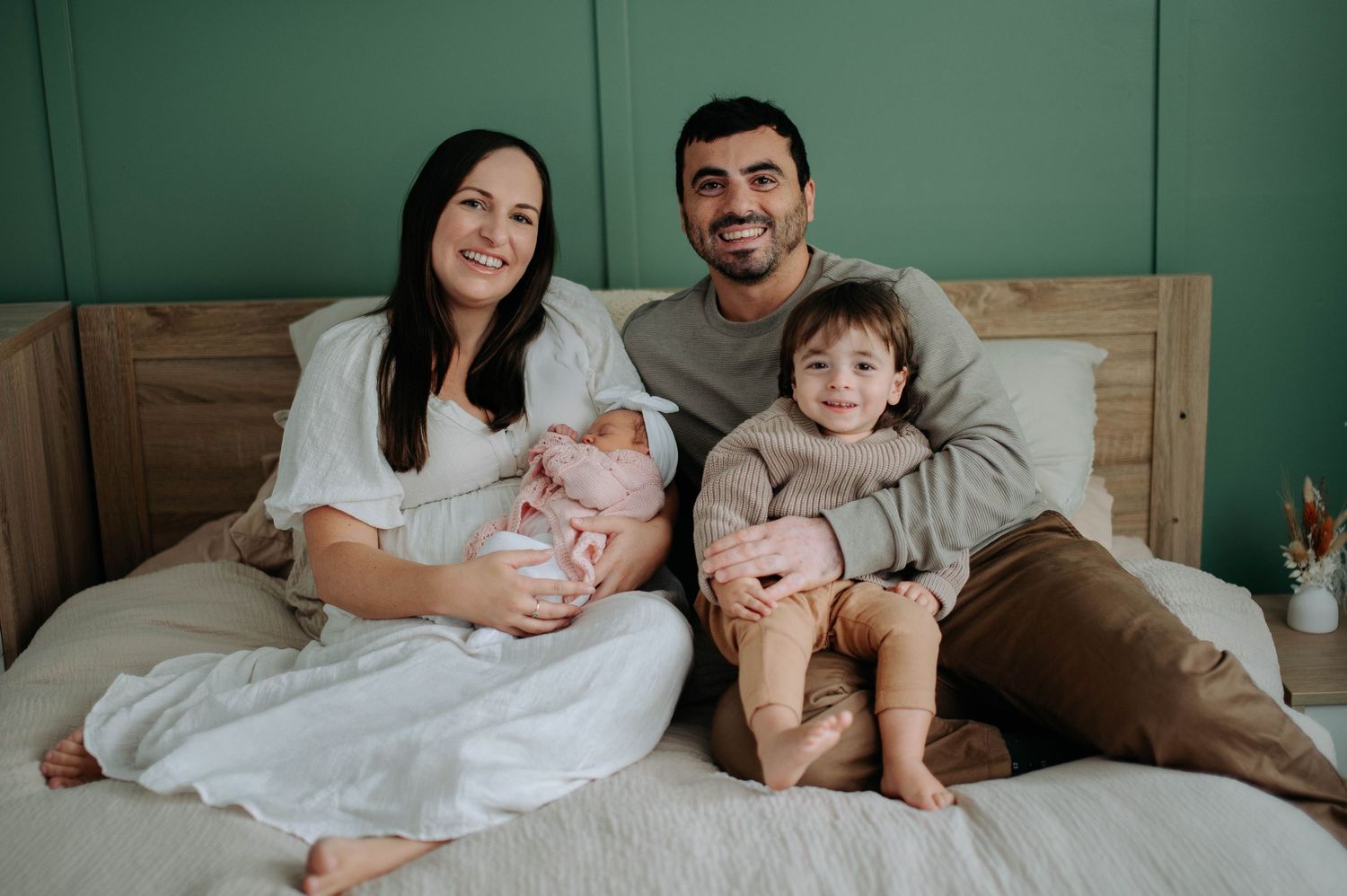 A family sits together on a bed with teal wall background in a cozy lifestyle portrait session at home.