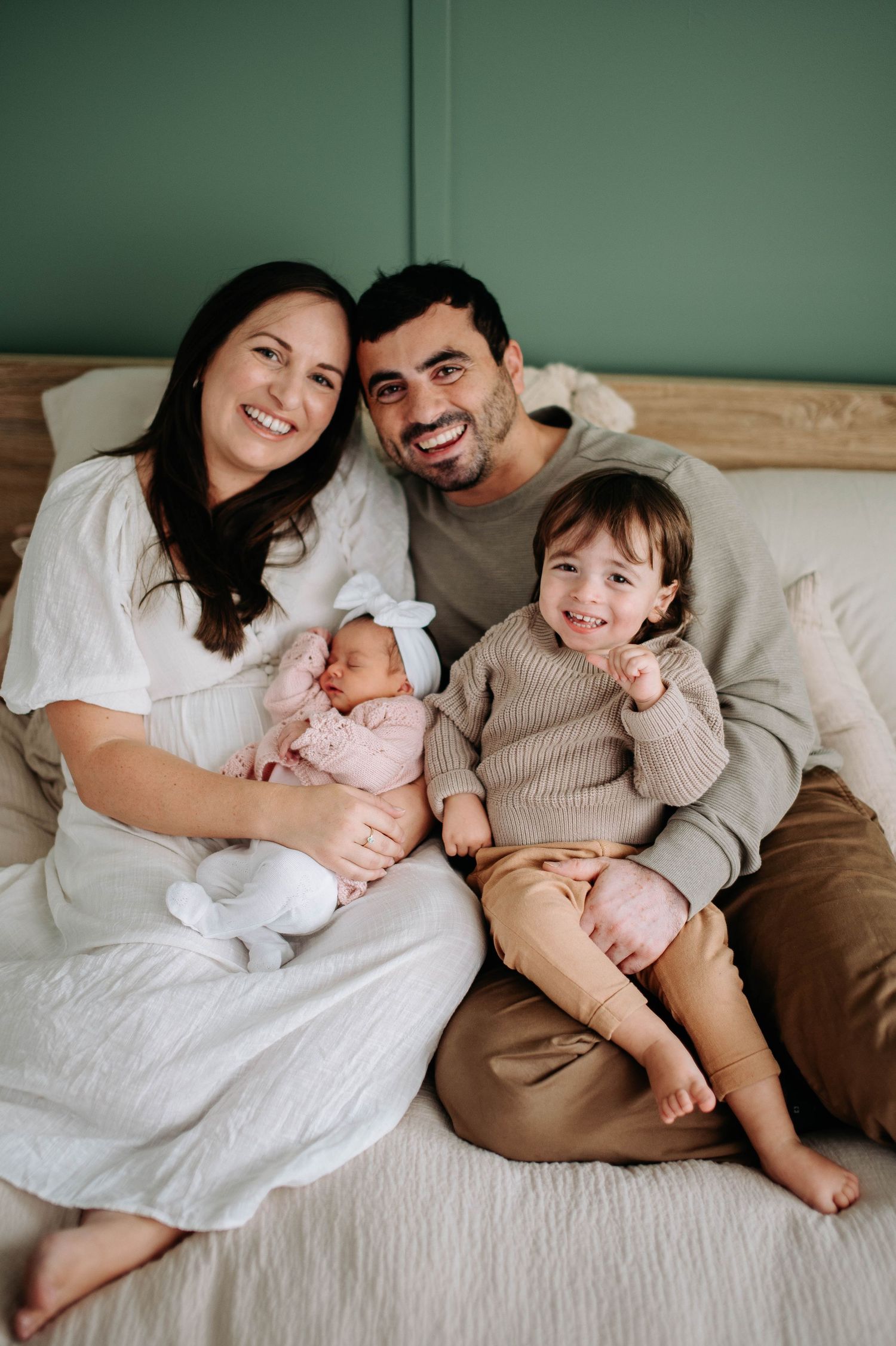 A family relaxing together on a bed with a newborn baby and toddler in casual attire.