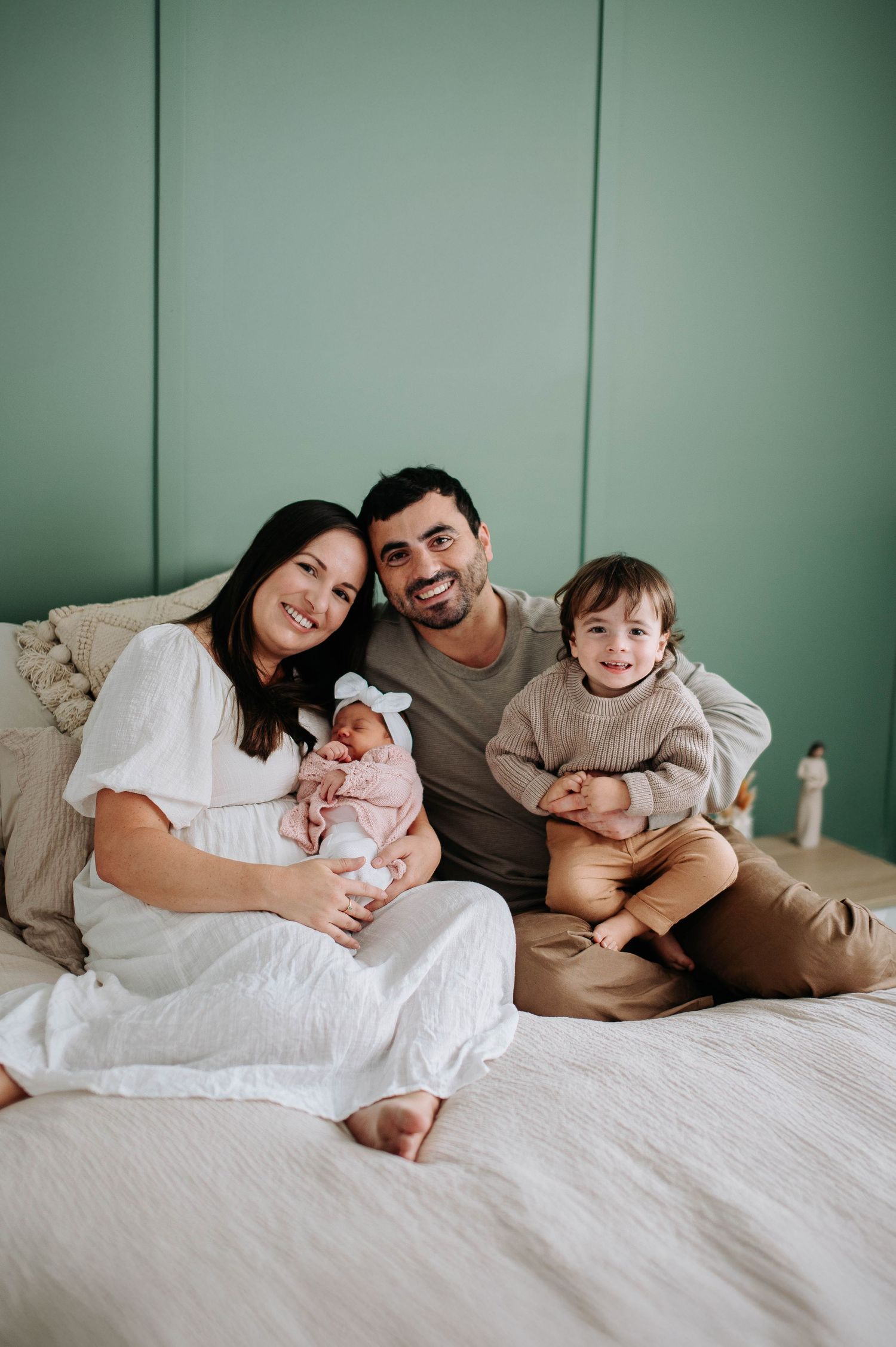 Family sits together on bed with newborn baby and toddler against mint green wall in lifestyle photo.
