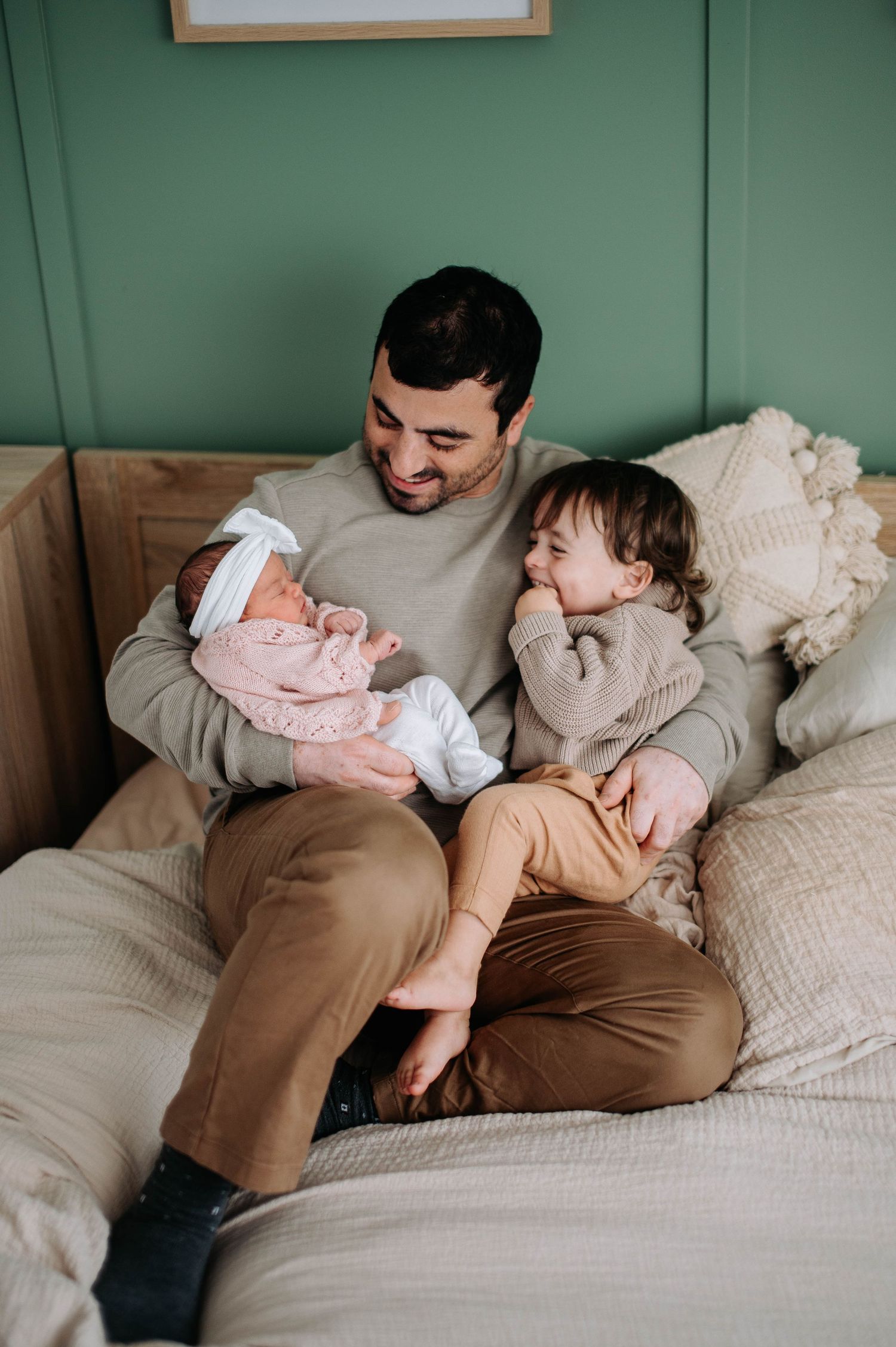 Family sharing tender moments with newborn baby on cozy bed with green wall and white linens in background.