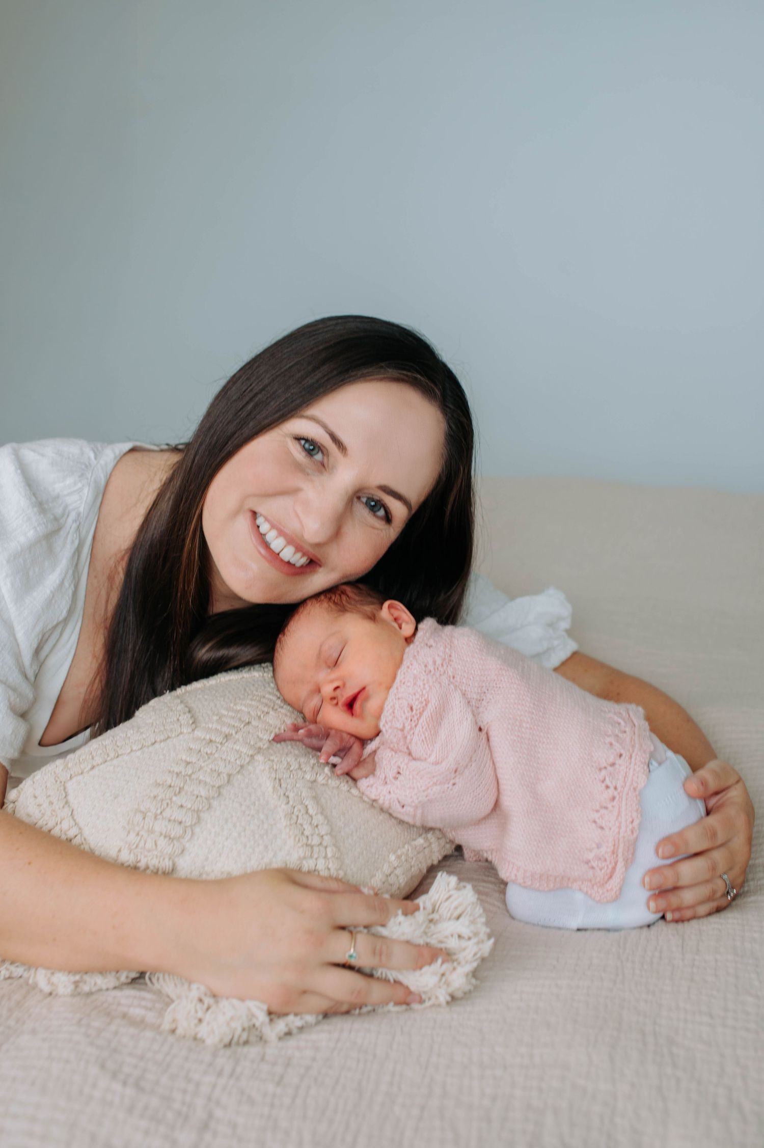 Mother and newborn baby in pink outfit lying together on beige couch sharing tender moment at home.