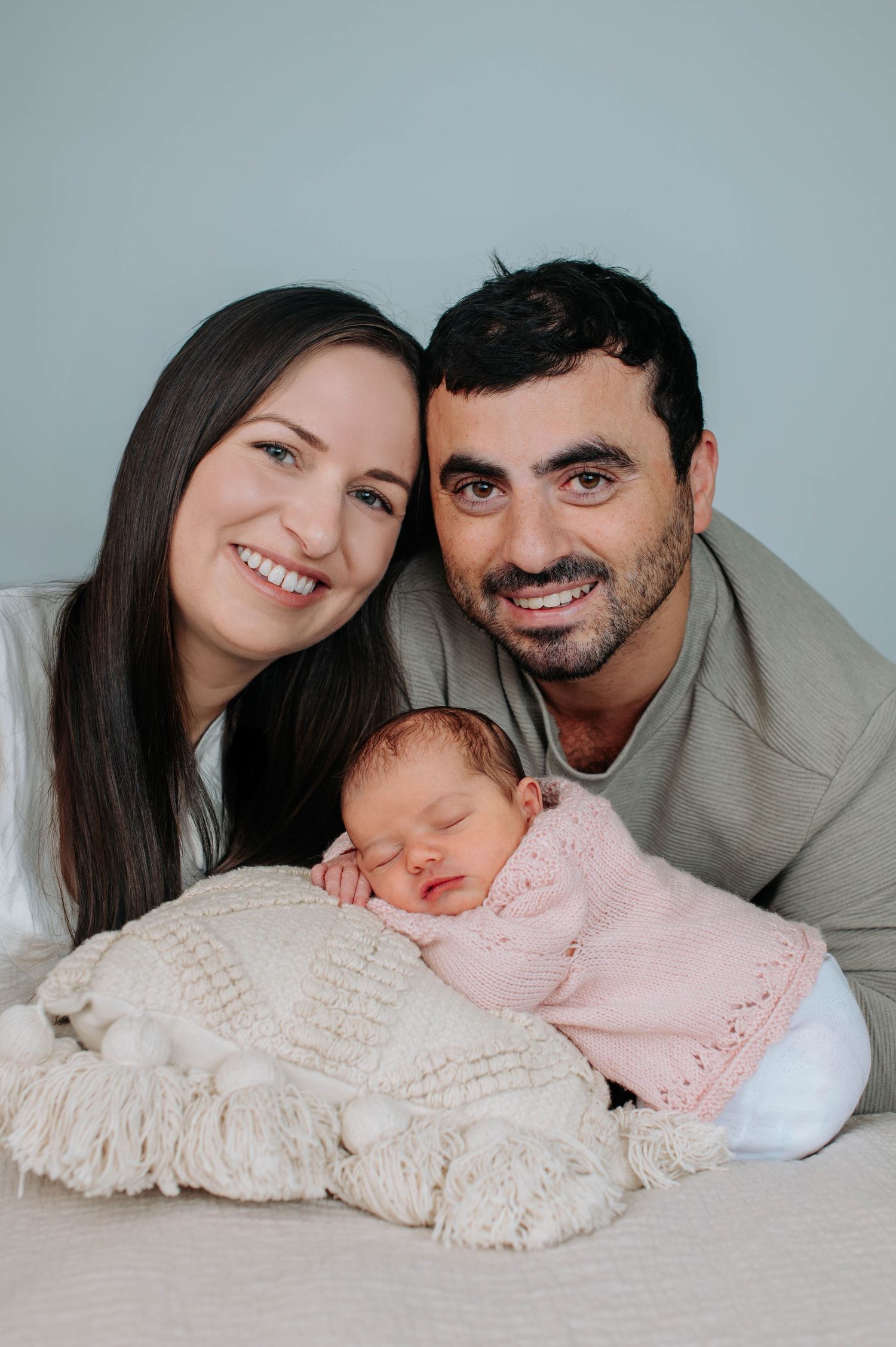 A sleeping newborn in pink outfit lies on cream blanket while parents lovingly watch from above in studio setting.
