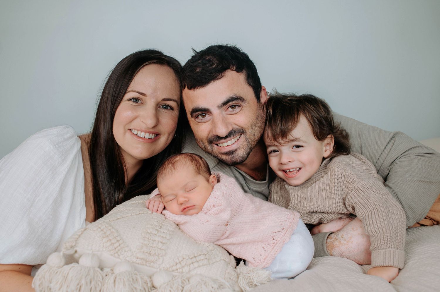 A loving family poses together on a bed, featuring a newborn baby wrapped in pink and a toddler in beige knitwear.