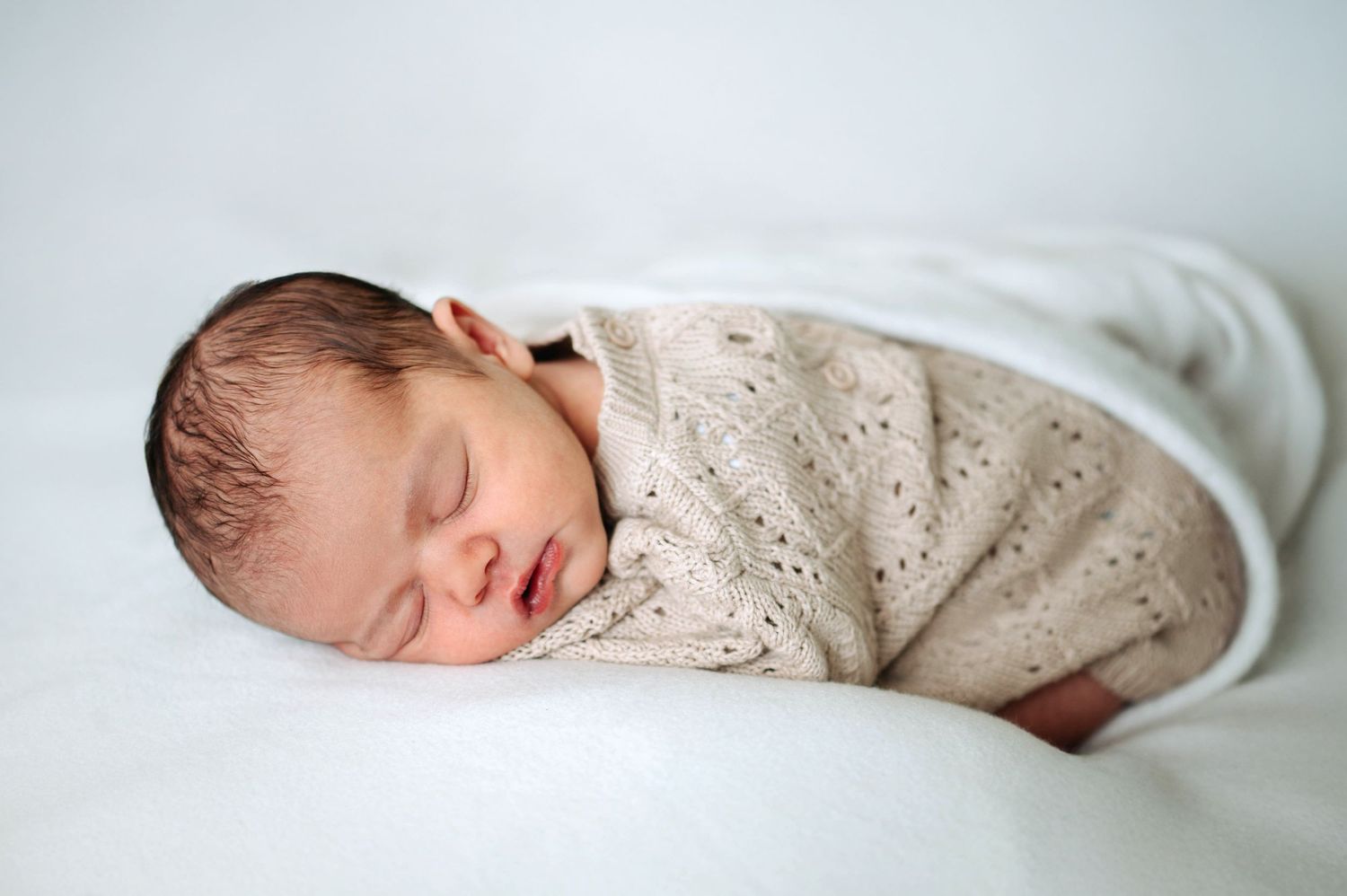 Sleeping newborn baby swaddled in white blanket and wearing cream knit sweater rests peacefully on light background.