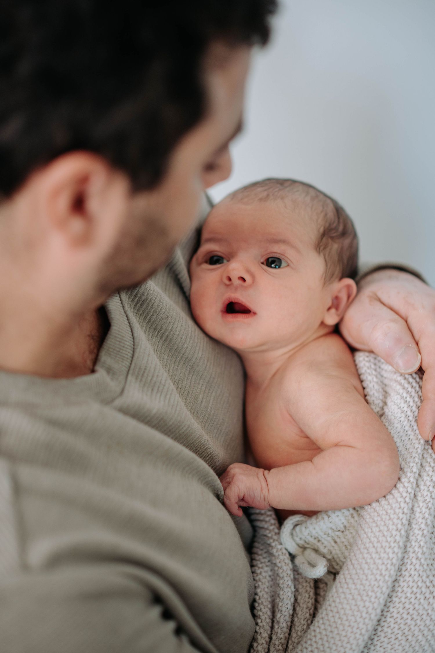 A tender moment shows a parent in gray holding a small baby swaddled in white knit fabric.
