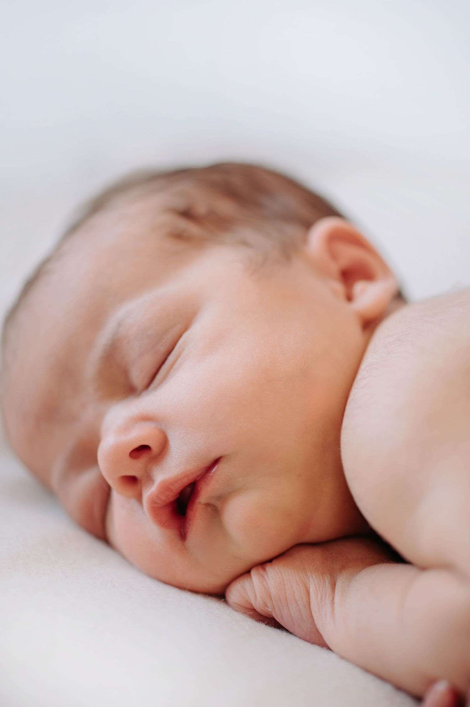 A sleeping newborn baby rests peacefully with soft rosy skin against white bedding in a closeup portrait.