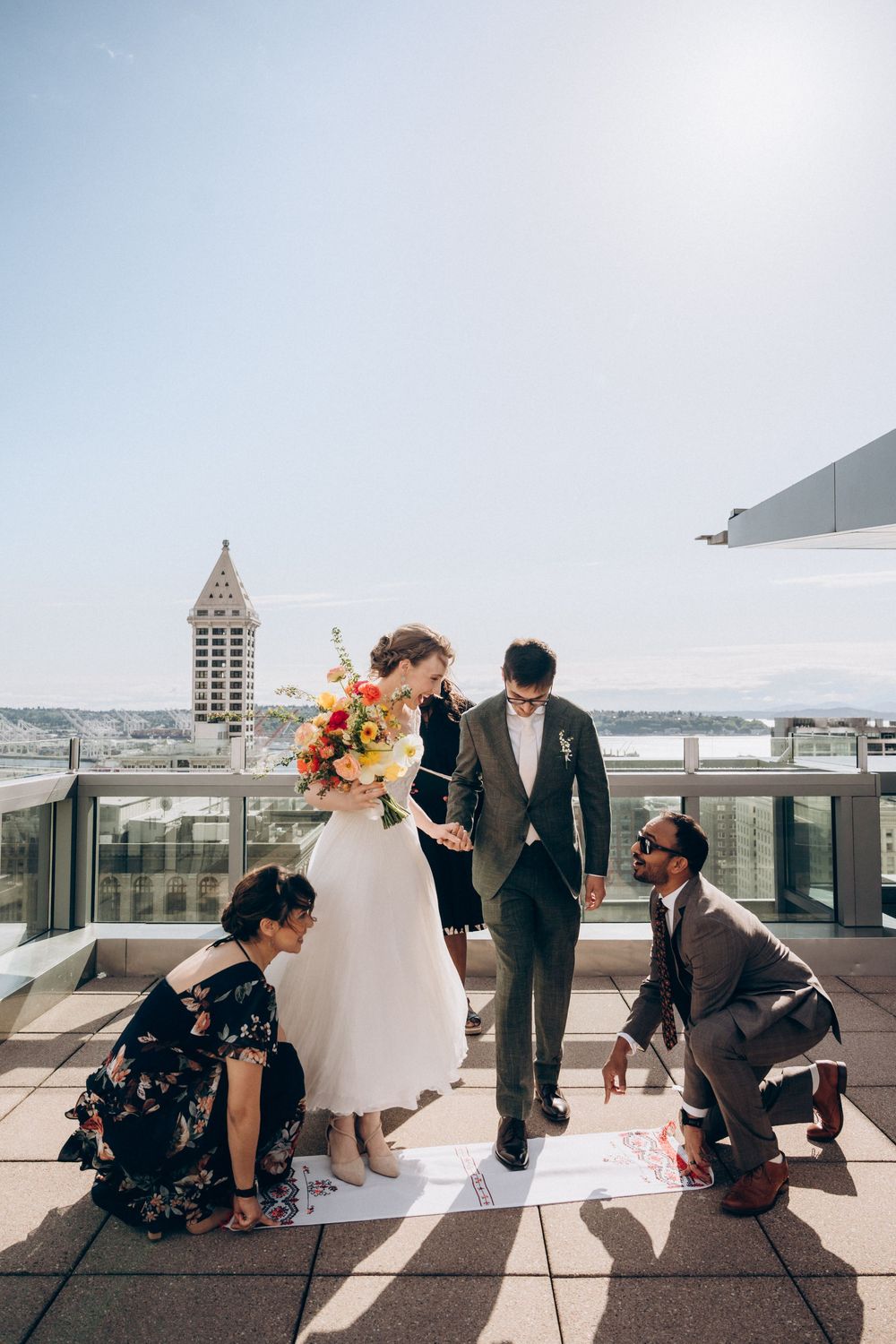Wedding party members help with Ukrainian wedding tradition called "Rushnyk"  on a rooftop terrace with the Smith Tower in Seattle backdrop.