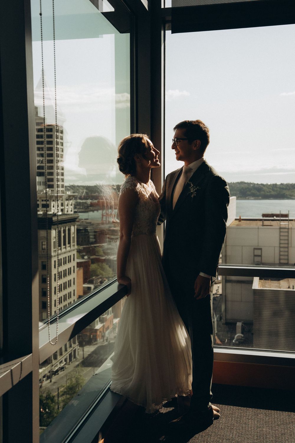 Silhouetted couple stands by large window in wedding attire overlooking city skyline at sunset.