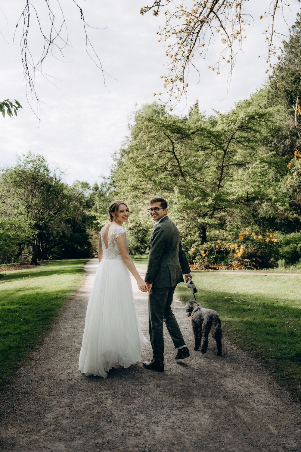 Two people in wedding attire walk along a park path as their dog accompanies them on an summer day.