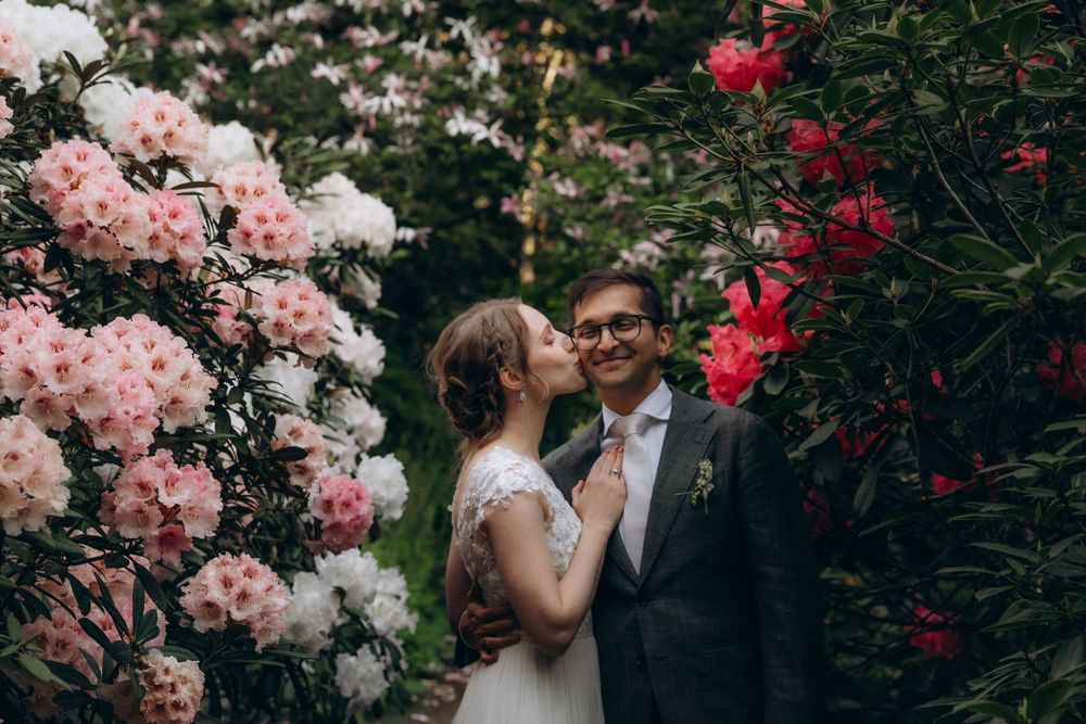 A romantic wedding photo featuring couple posing amid blooming pink and red azalea bushes in a garden setting.