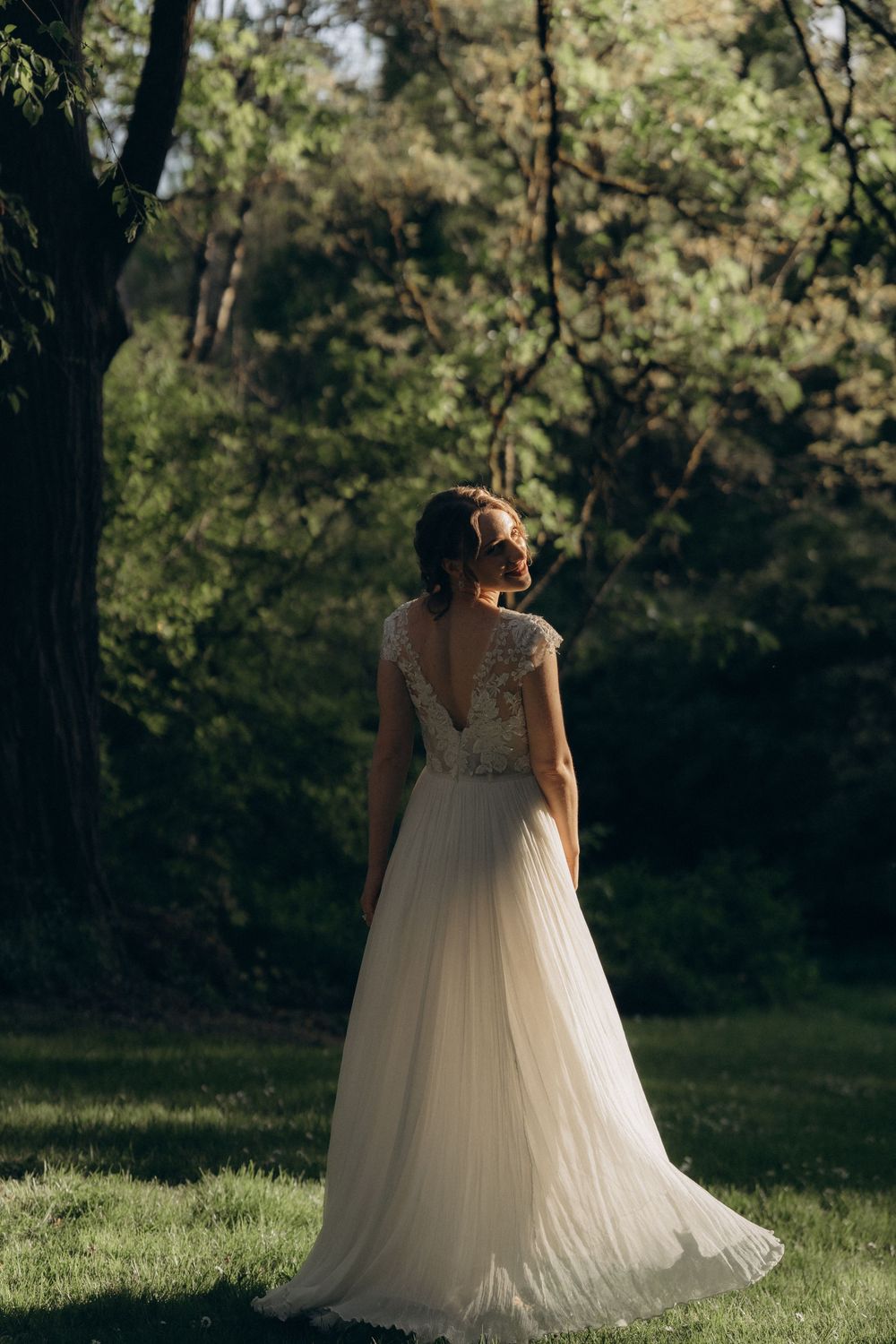 A bride in a flowing white backless gown with lace detail stands in a sunlit garden setting among trees.