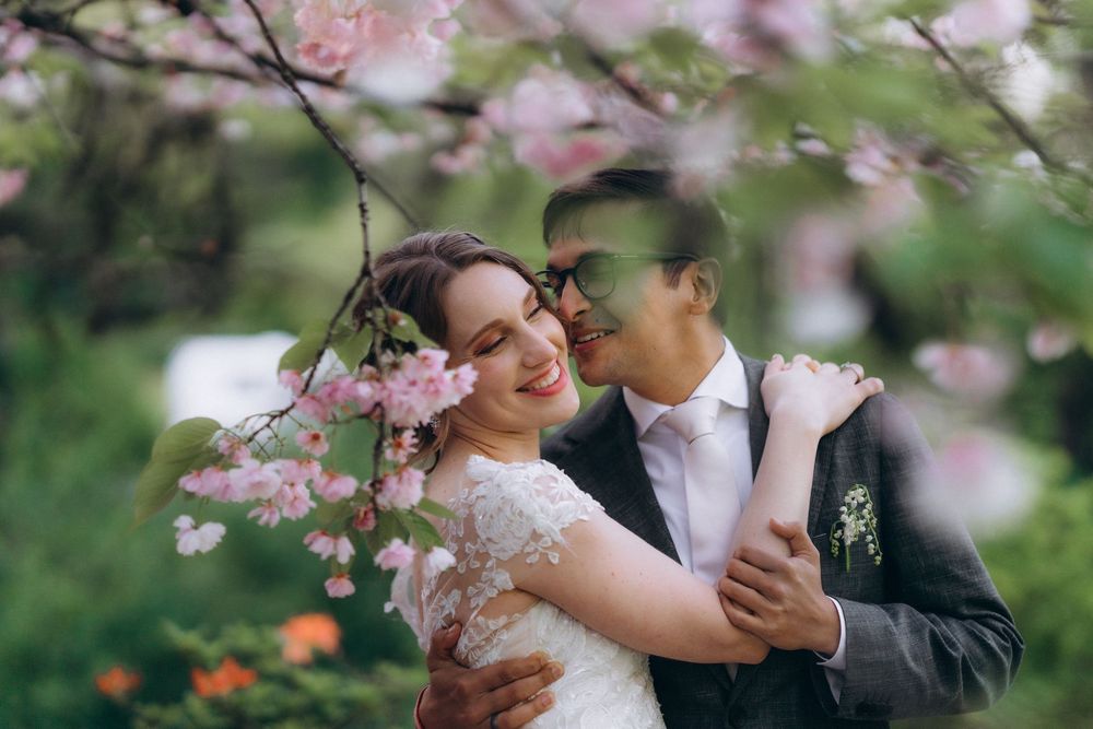 Couple embraces among delicate pink cherry blossoms in a romantic spring garden setting.