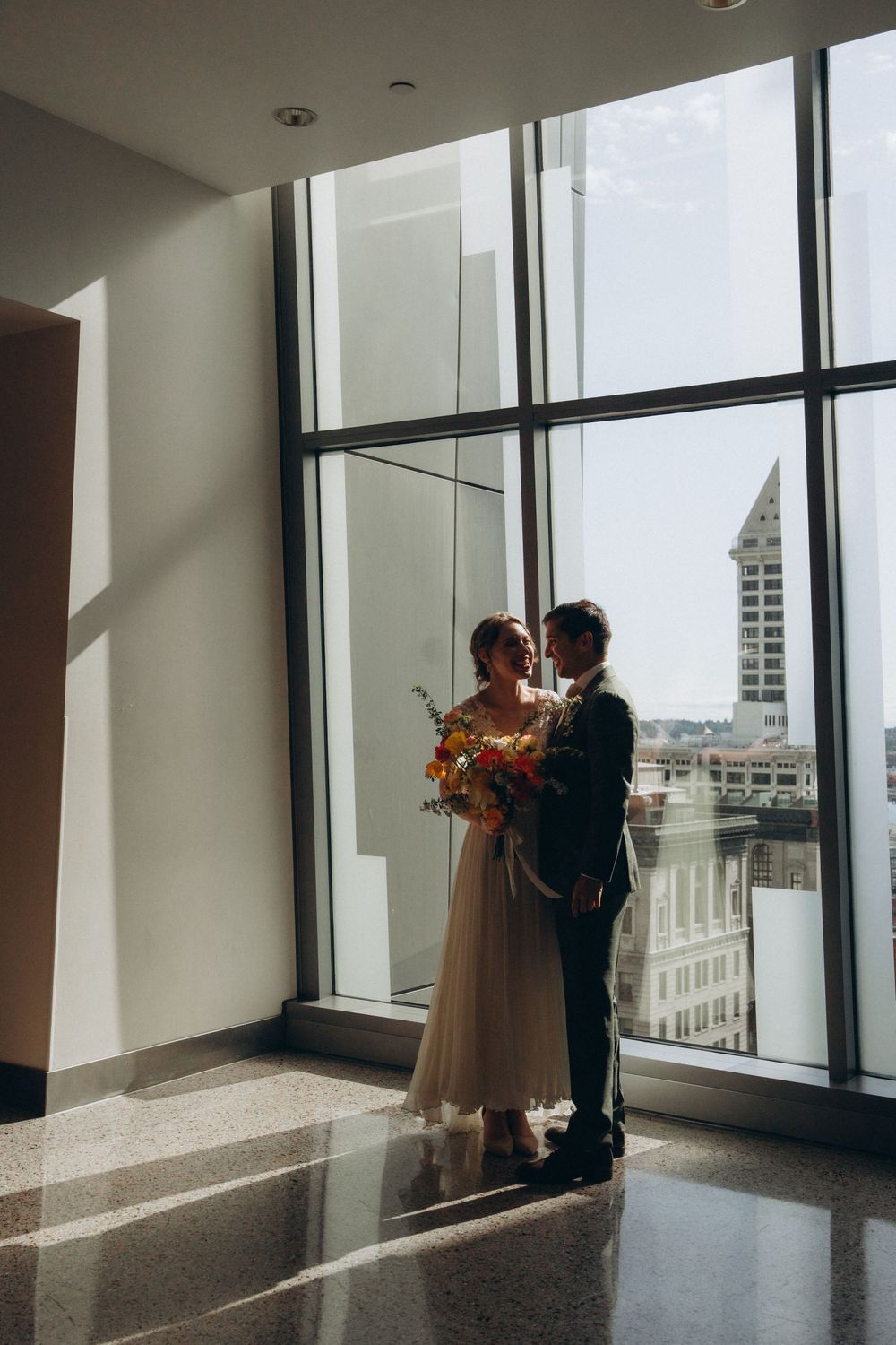 A romantic wedding moment silhouetted against large windows overlooking the Seattle Smith Tower and city skyline.