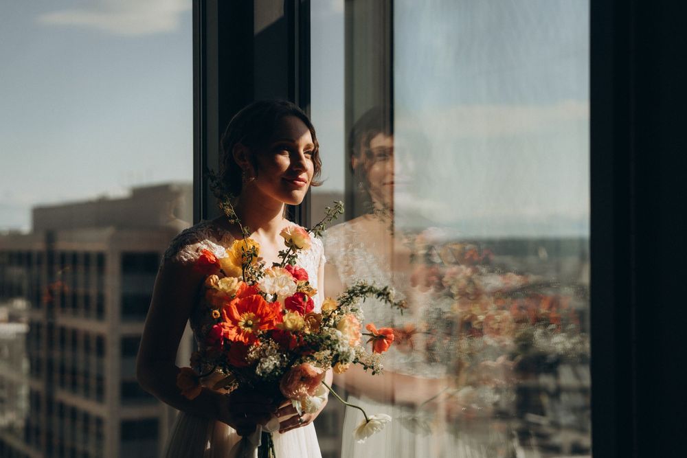 Wedding photographer captures silhouette of bride with sunset bouquet against urban cityscape through window reflection.
