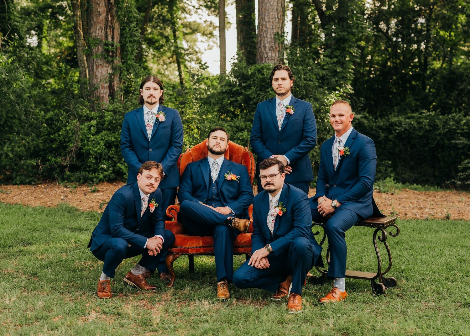 Groomsmen in matching navy suits pose together on garden furniture surrounded by tall trees and green grass in East Texas.