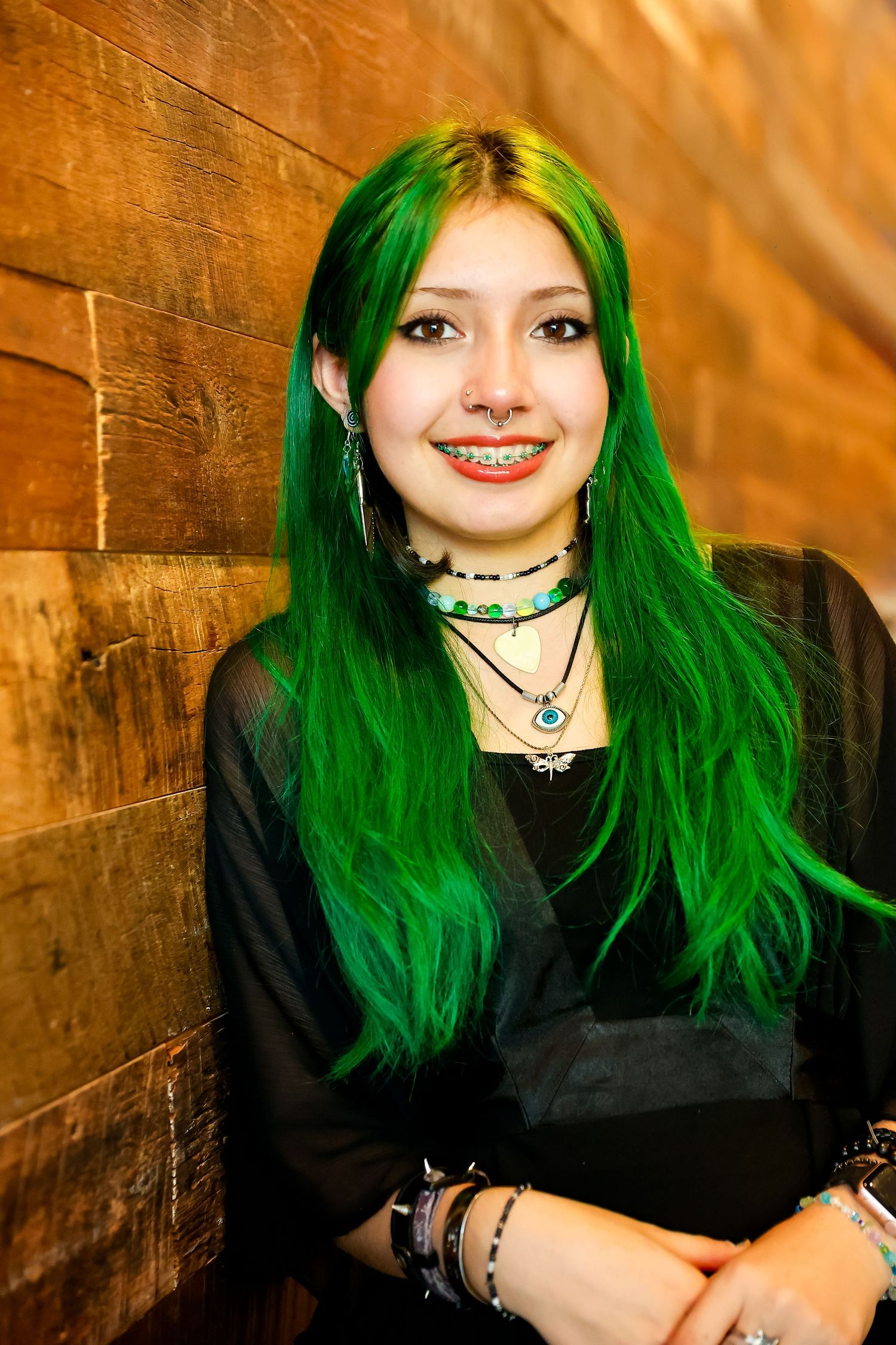 A figure in a black top poses against a rustic wooden wall with warm lighting.