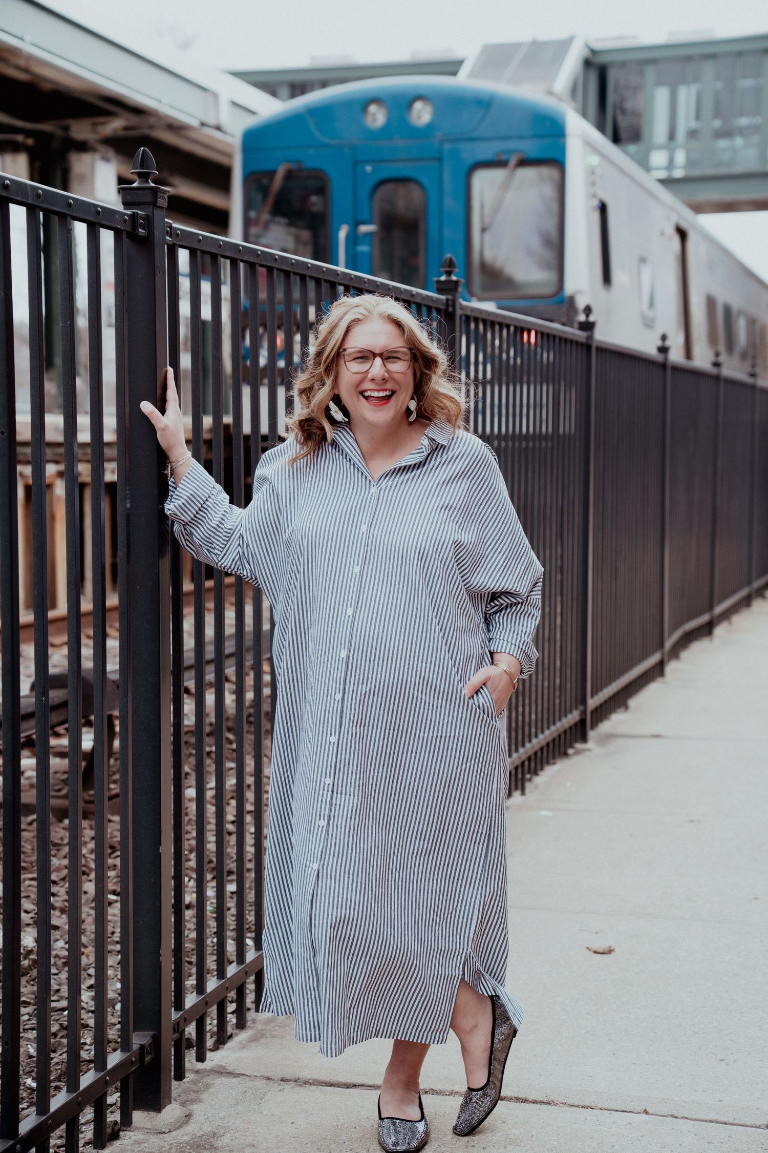 A person in a striped shirt dress poses cheerfully along an urban railing.