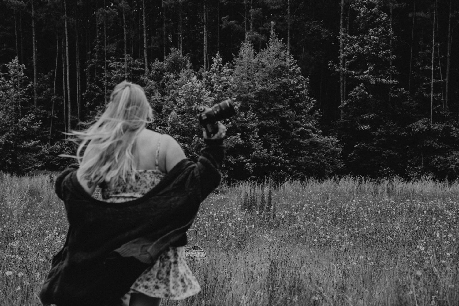 A figure stands in a wildflower meadow at the edge of a dark forest, wearing a floral dress and twirling.