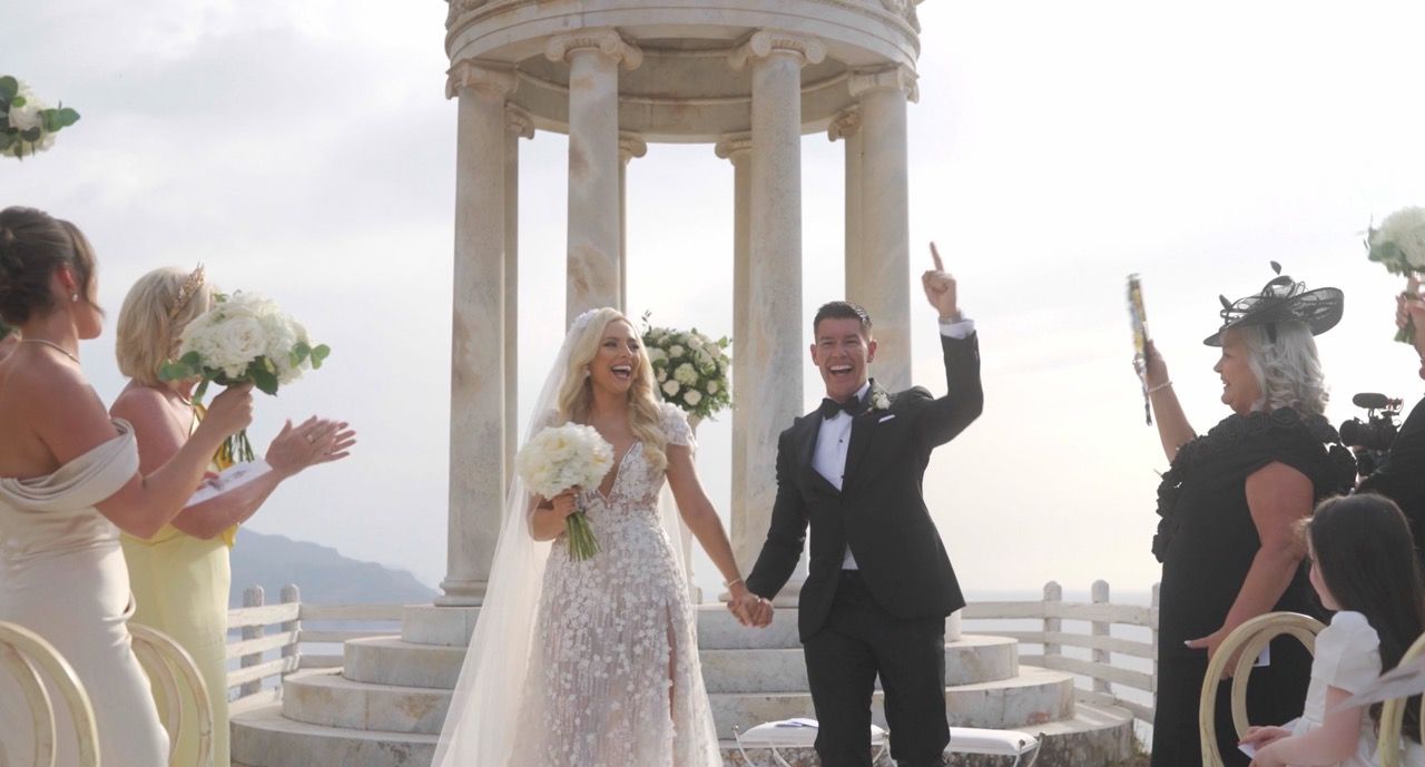 Bride and groom celebrating their wedding in an outdoor ceremony, surrounded by guests and a scenic backdrop with a classical gazebo.