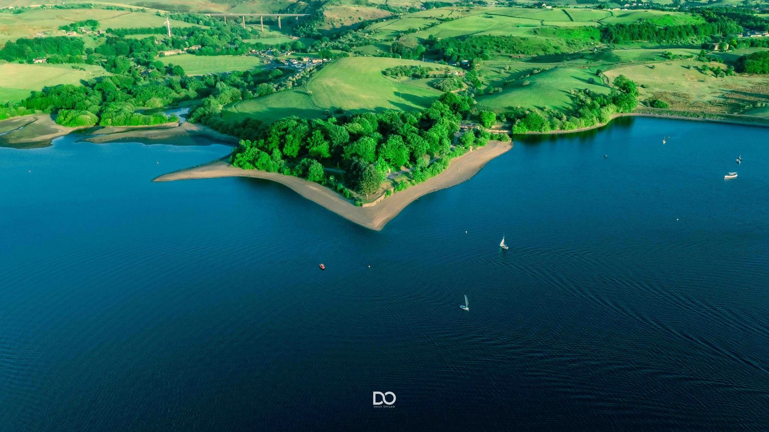 Aerial view of a lush green peninsula with sailboats dotting the deep blue waters of a lake or reservoir.