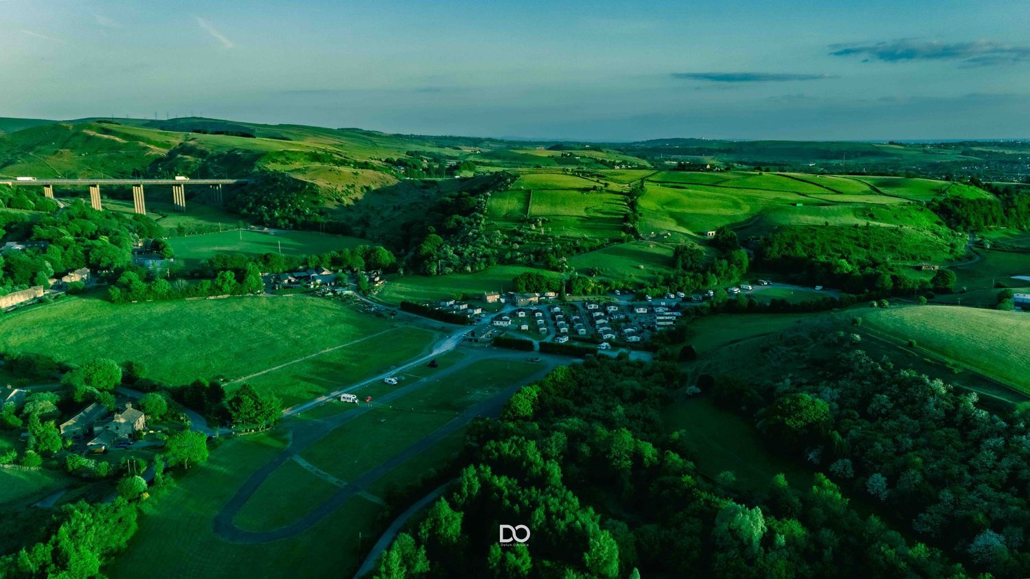 Aerial view of a green rural landscape with a campground nestled among rolling hills and a bridge in the distance.