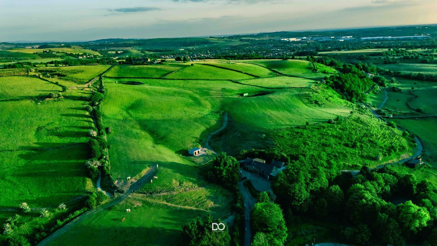 Aerial view of lush green rolling hills with winding river and patches of forest under cloudy skies.