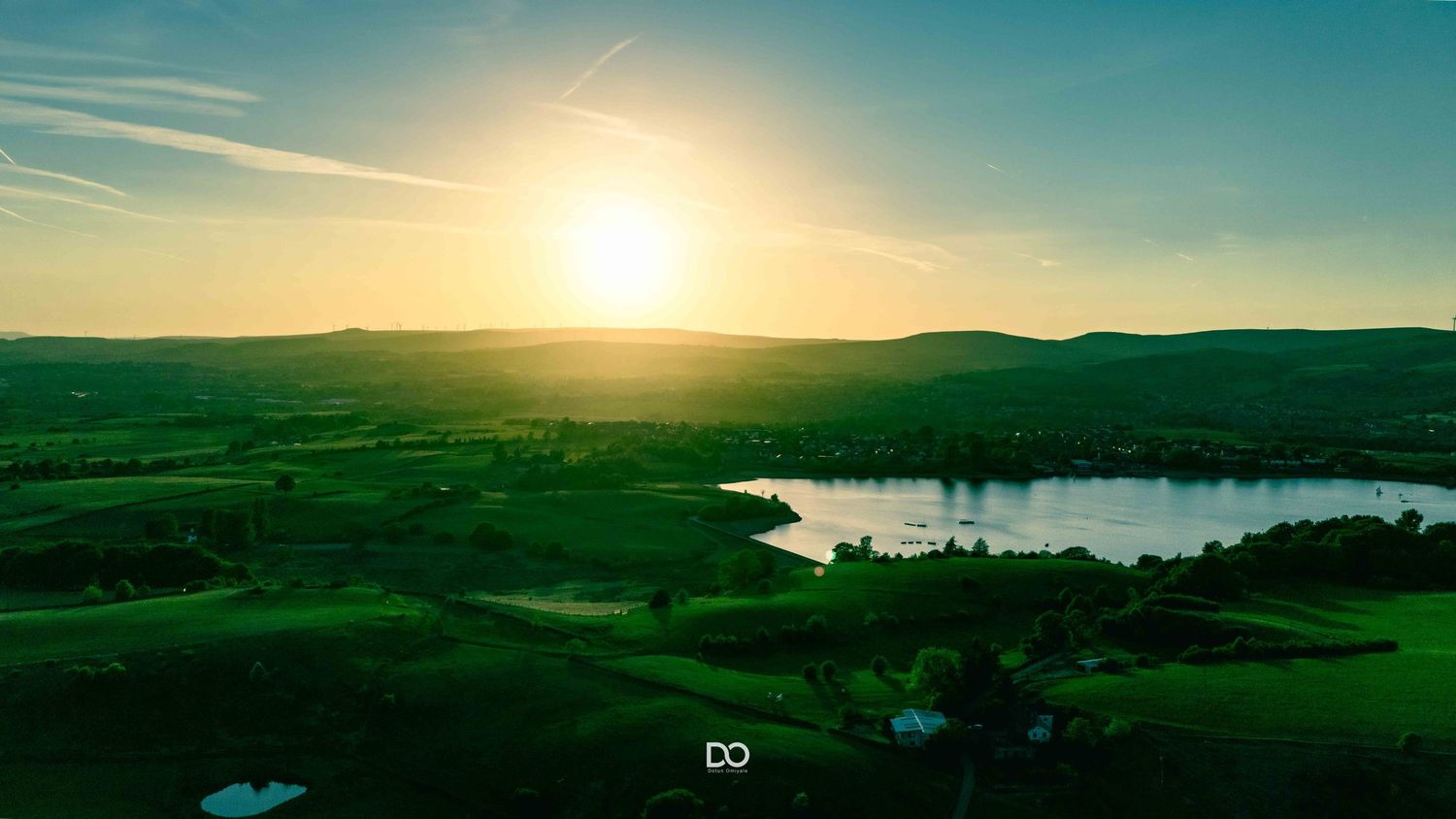 Rolling green hillsides stretch out beneath a brilliant sunset sky with lakes and mountains in the panoramic landscape.