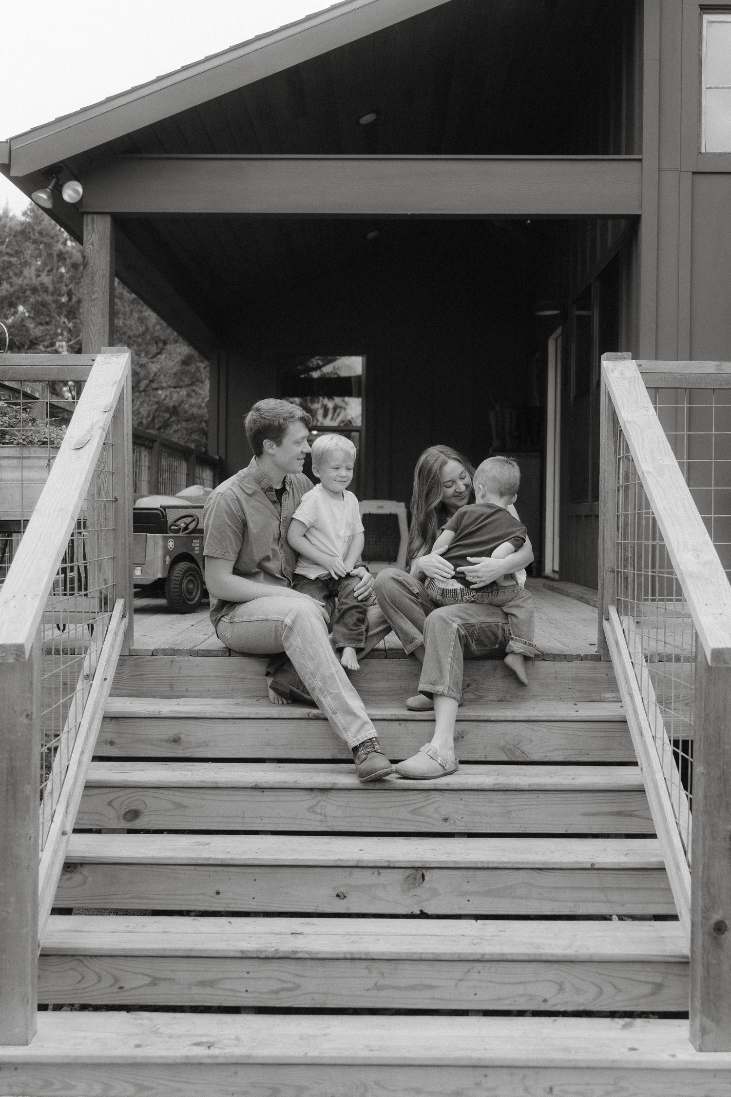 A family sits together on wooden stairs of a house entrance in a casual black and white photograph.