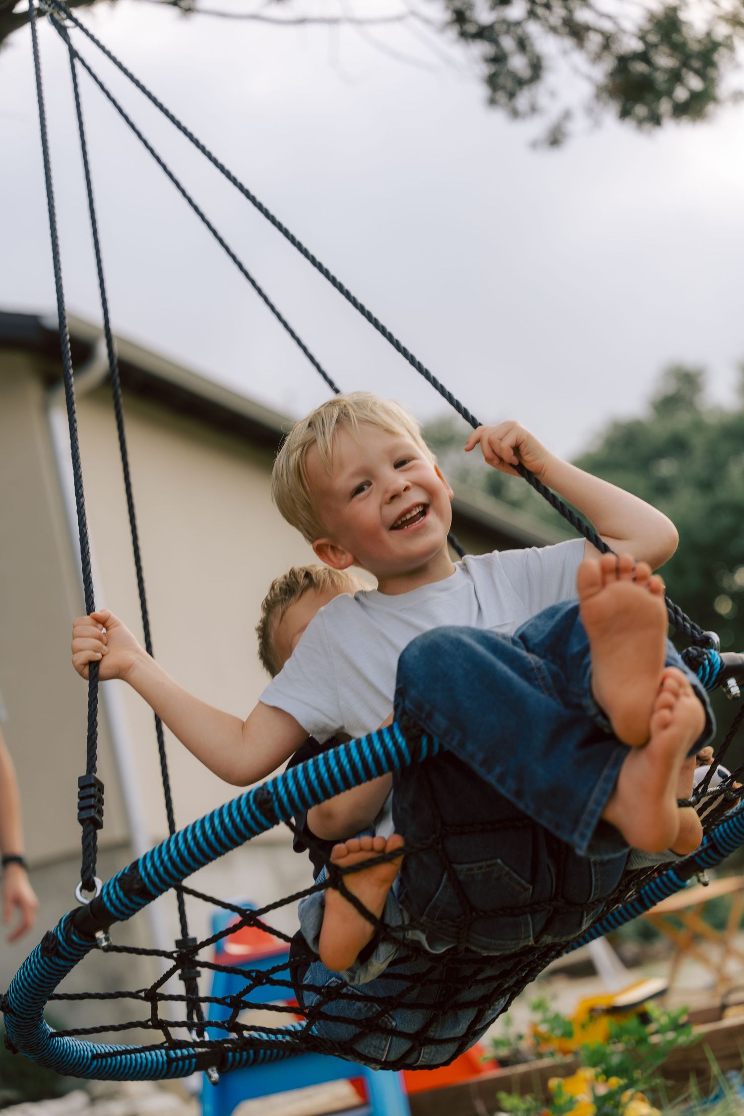 A joyful young child swings on a large disc-shaped outdoor swing with bare feet against a cloudy sky.
