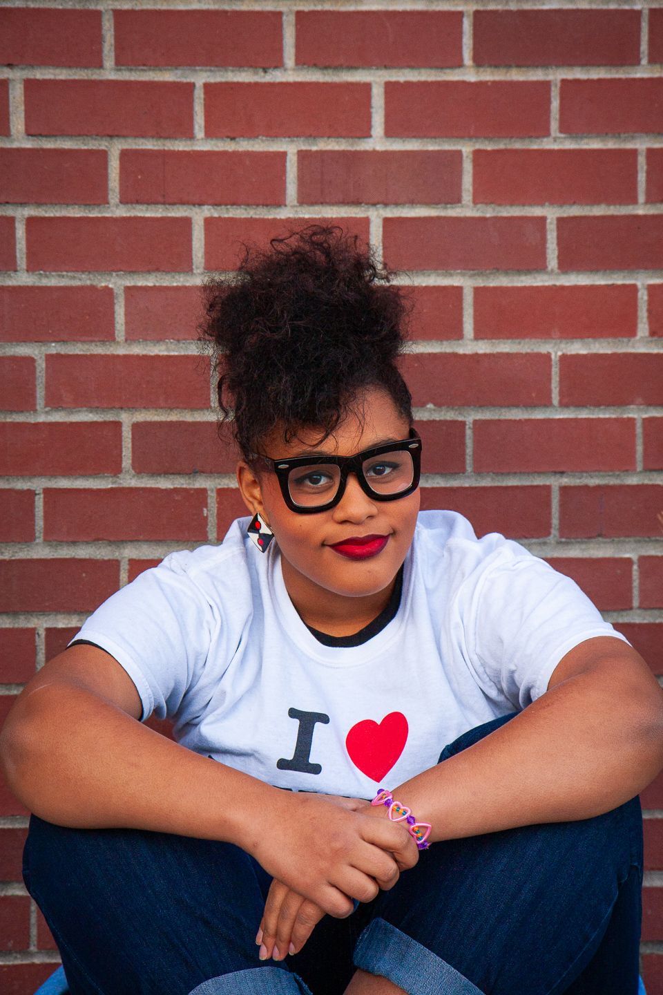Someone wearing glasses and a white 'I heart' t-shirt sits against a red brick wall with a bright smile.