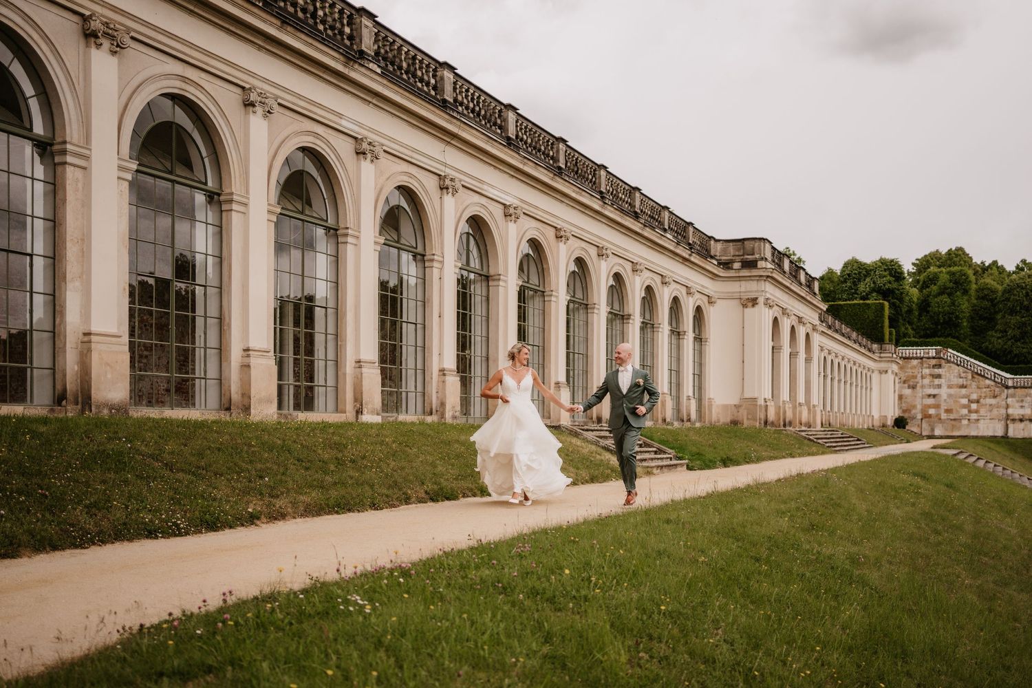Two figures run along a path beside a grand neoclassical colonnade with arched windows at a historic estate.