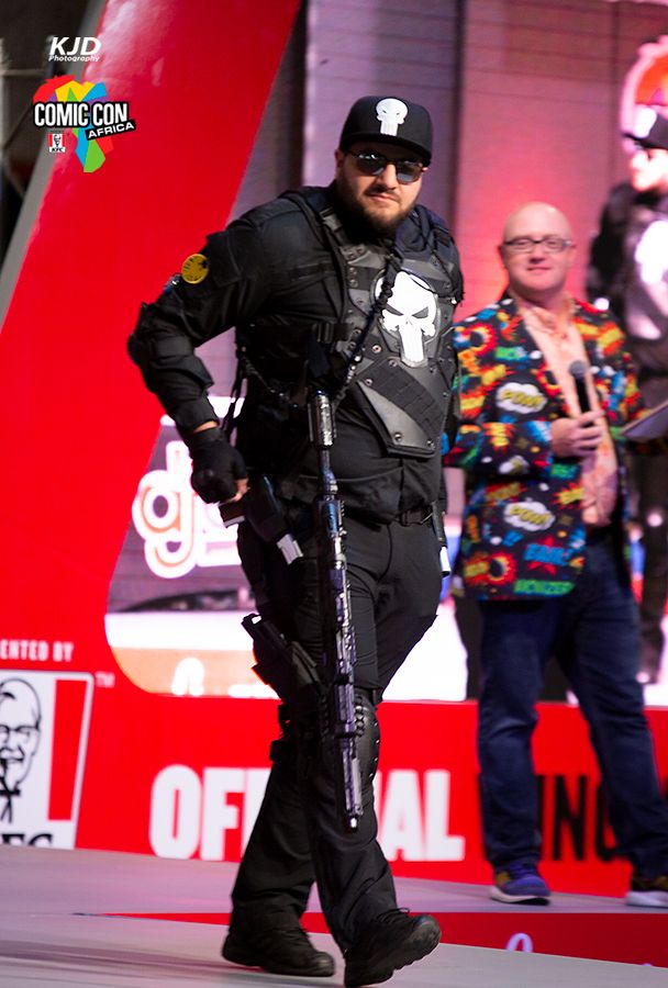 Security officer in black tactical gear crouches with weapon drawn at Comic Con event with red backdrop and crowd visible.