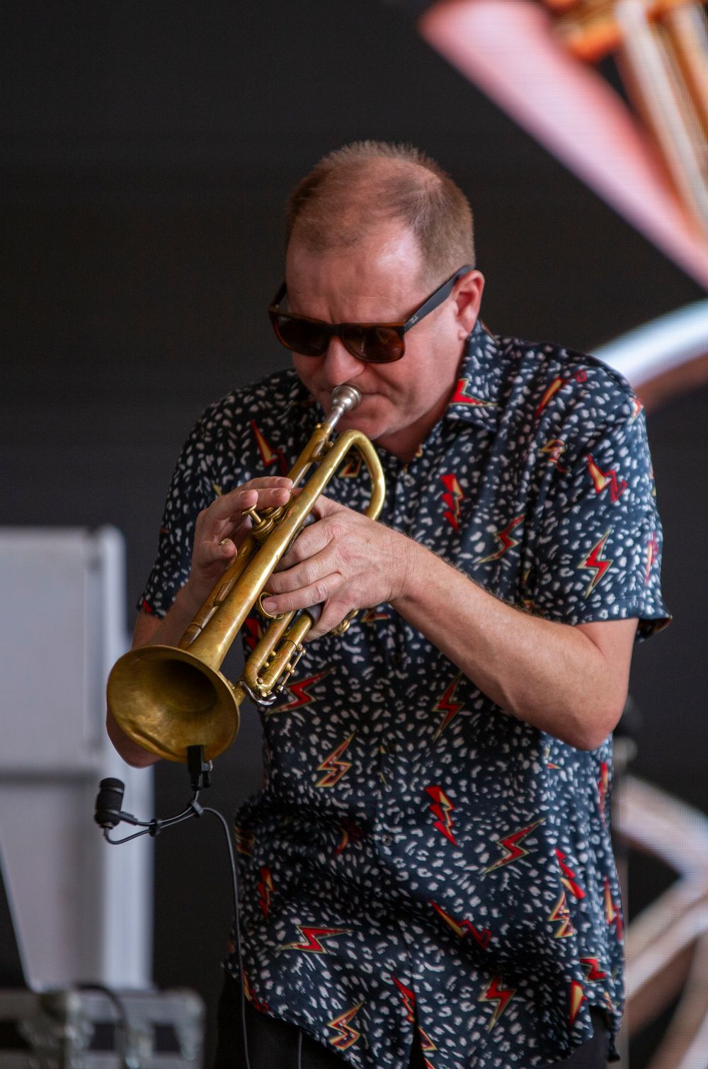 Jazz musician plays a brass trumpet while wearing sunglasses and a patterned short-sleeve shirt during a performance.