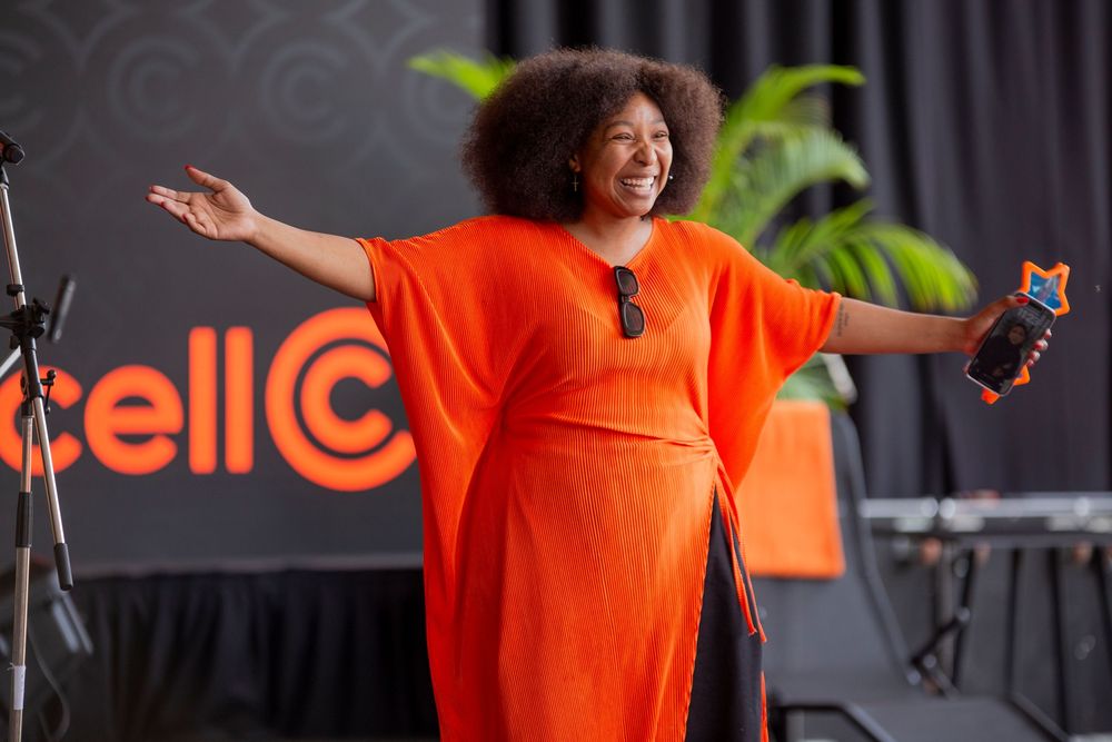 A performer in a bright orange dress gestures enthusiastically on stage with arms outstretched against an orange backdrop.