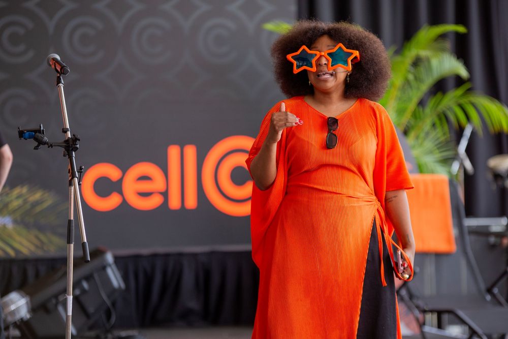 A performer in a bright orange dress and star-shaped glasses stands on stage with the Cell C logo visible in the background.