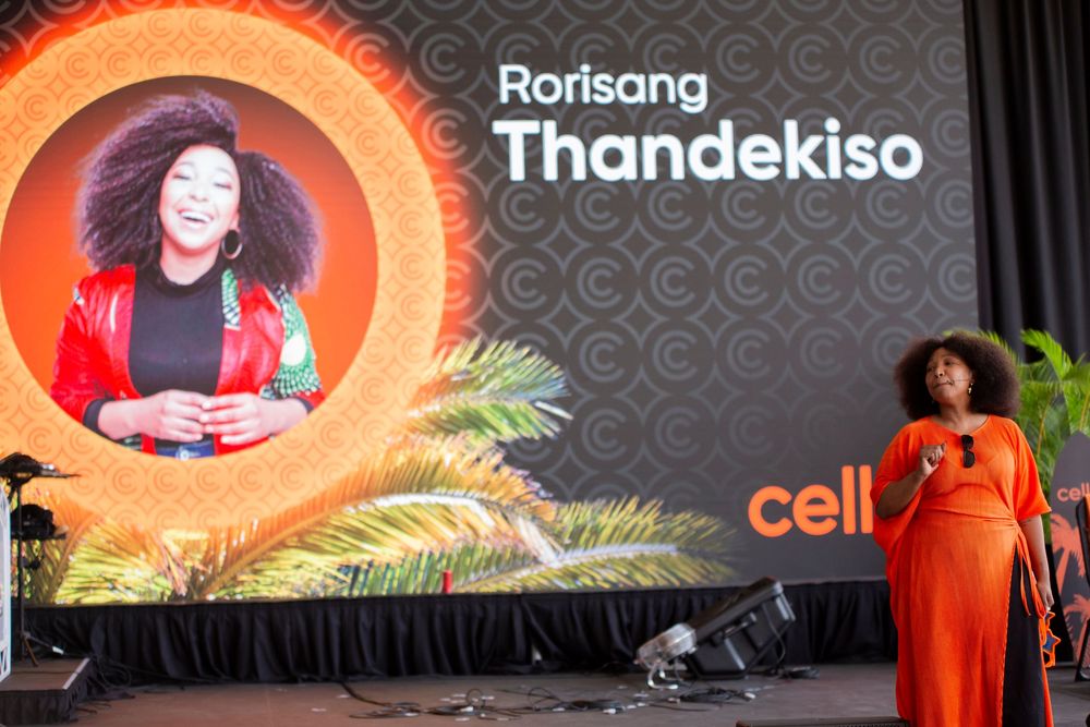 A performer in a bright orange dress stands on stage with a circular branded backdrop display at an event.