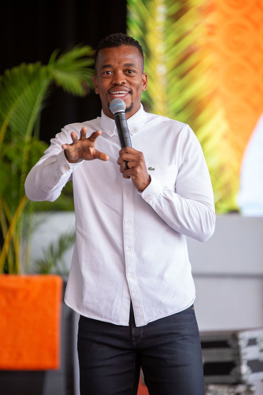 A performer speaks into a microphone while wearing a white button-down shirt and dark pants on stage with palm trees behind.