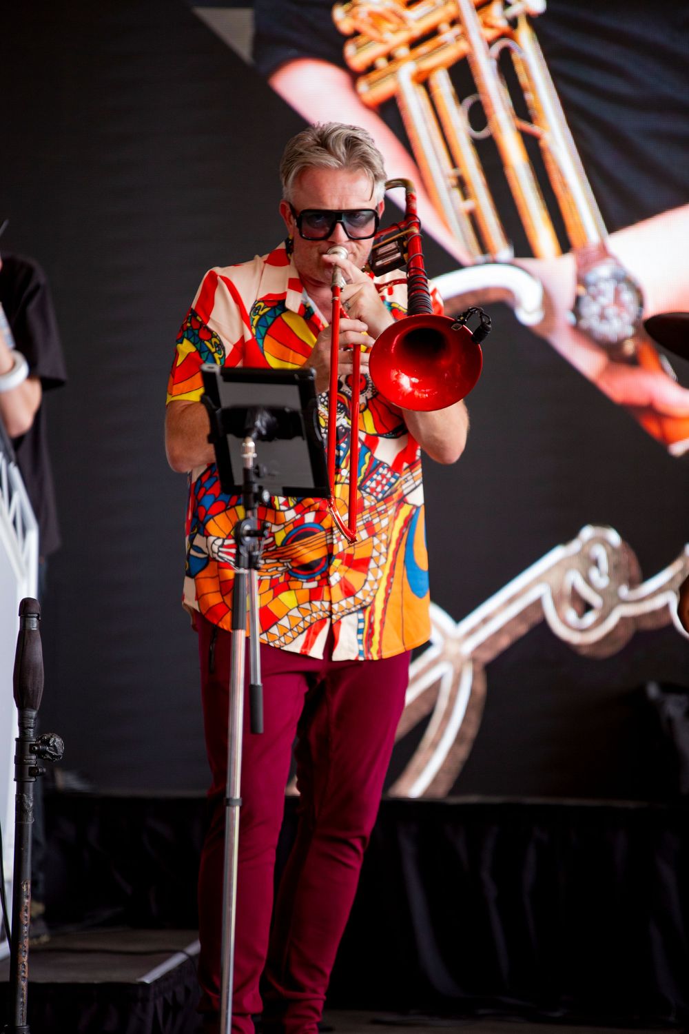 Musician in colorful patterned shirt and burgundy pants performs with a trumpet on stage against dark backdrop and lighting.
