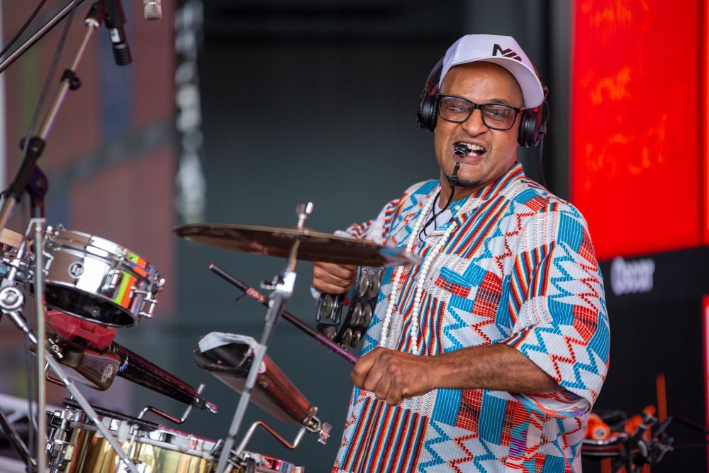 An enthusiastic drummer performs on stage wearing glasses, a baseball cap and a colorful patterned shirt.