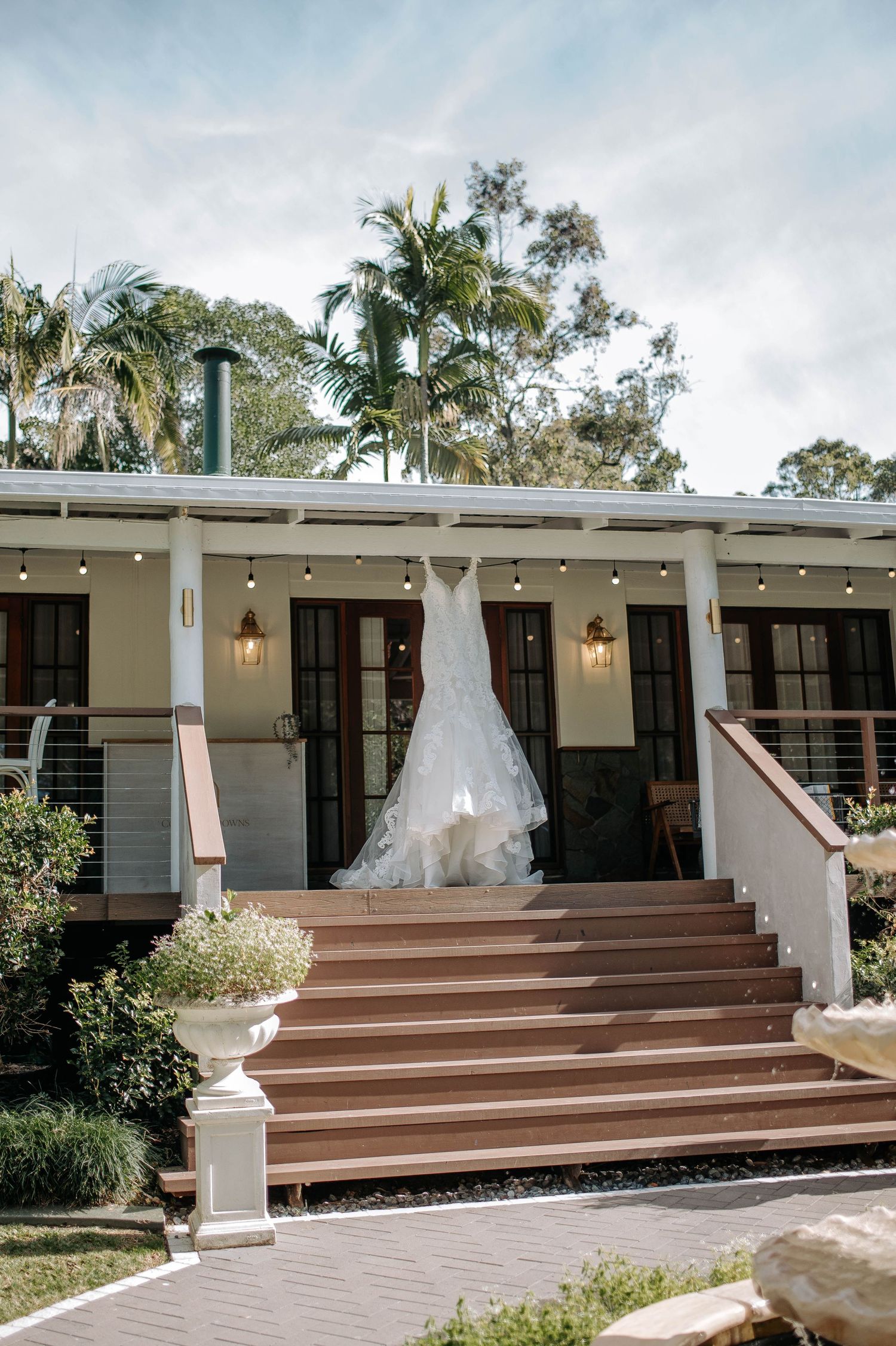 White wedding dress displayed on verandah of modern home with wood stairs and palm trees in tropical setting.