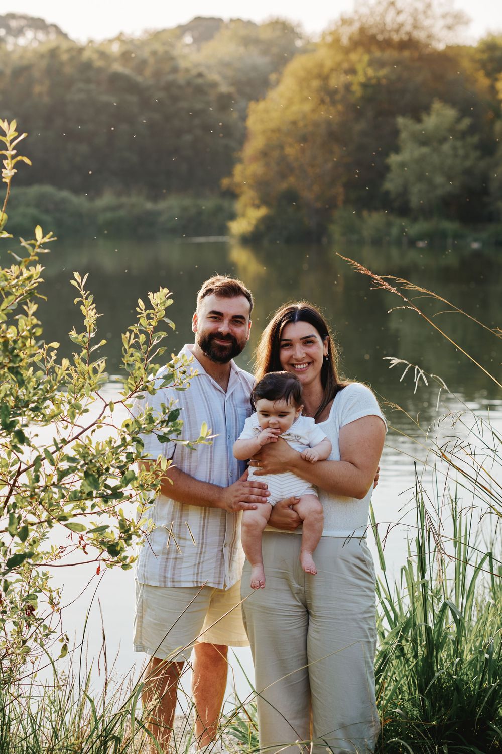 Smiling young family holding a baby standing near a sunlit lake with lush green trees in background.