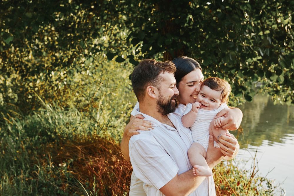 Happy young family with a baby sharing a tender moment outdoors near a lake surrounded by greenery.