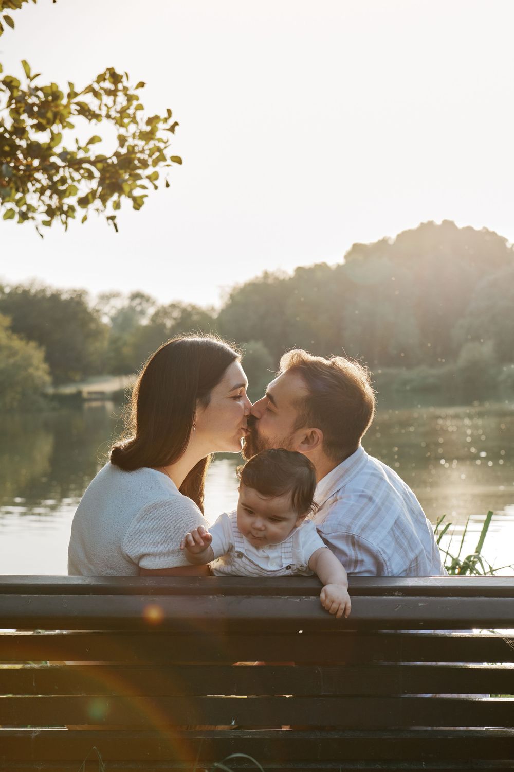 Couple sharing a romantic kiss with baby between them on a sunny lakeside bench.