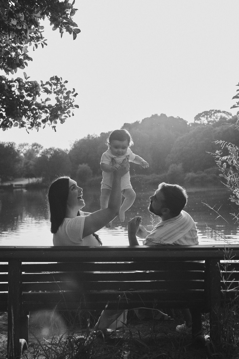 Black and white photo of parents lifting baby on a lakeside bench at sunset.